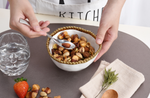 A person in a KITCHEN apron uses a spoon to serve an almond from a gold-rimmed white bowl of mixed nuts including pistachios and broad beans, with a glass of water and a red strawberry on a brown placemat