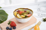 A colorful fruit salad in a white bowl with a gold beaded rim featuring red strawberries, green grapes, and yellow lemon slices, elegantly displayed on a light wooden cutting board with additional blueberries, an orange wedge, and a gold fork on a white marble surface by a window