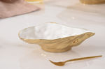 A decorative, irregularly shaped gold metallic serving bowl with a white interior and a matching gold appetizer fork rests on a reflective white marble surface with a soft pink napkin in the background