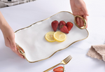 White wavy ceramic serving tray with gold handles and rim, held by a person, displaying fresh red strawberries and bright yellow lemon slices on a white marble surface with a gold fork and beige towel nearby