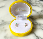 Product photo by Jack and Jewels of a pair of gold stud earrings featuring three tiny pastel pink mint green and light blue enamel stars displayed in an open round yellow velvet box on a white marble surface
