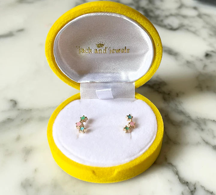 Product photo by Jack and Jewels of a pair of gold stud earrings featuring three tiny pastel pink mint green and light blue enamel stars displayed in an open round yellow velvet box on a white marble surface