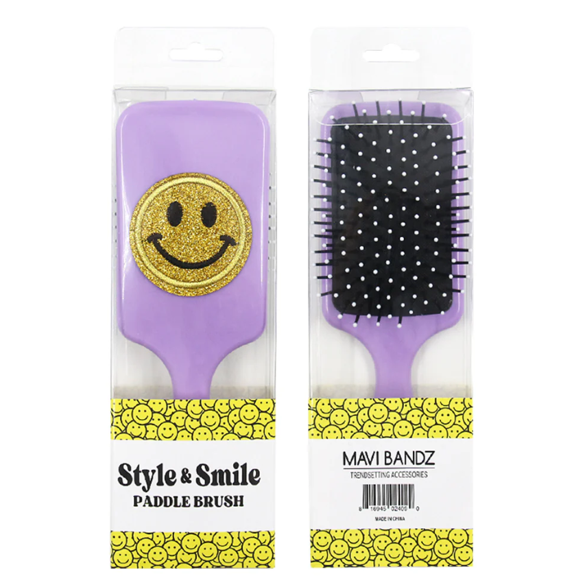 Product photography of two Mavi Bandz Style & Smile lavender paddle brushes in clear retail packaging, one adorned with a prominent gold glitter smiley face, the other displaying black bristles with white polka dot tips, resting on packaging featuring a yellow repeating smiley face pattern and text