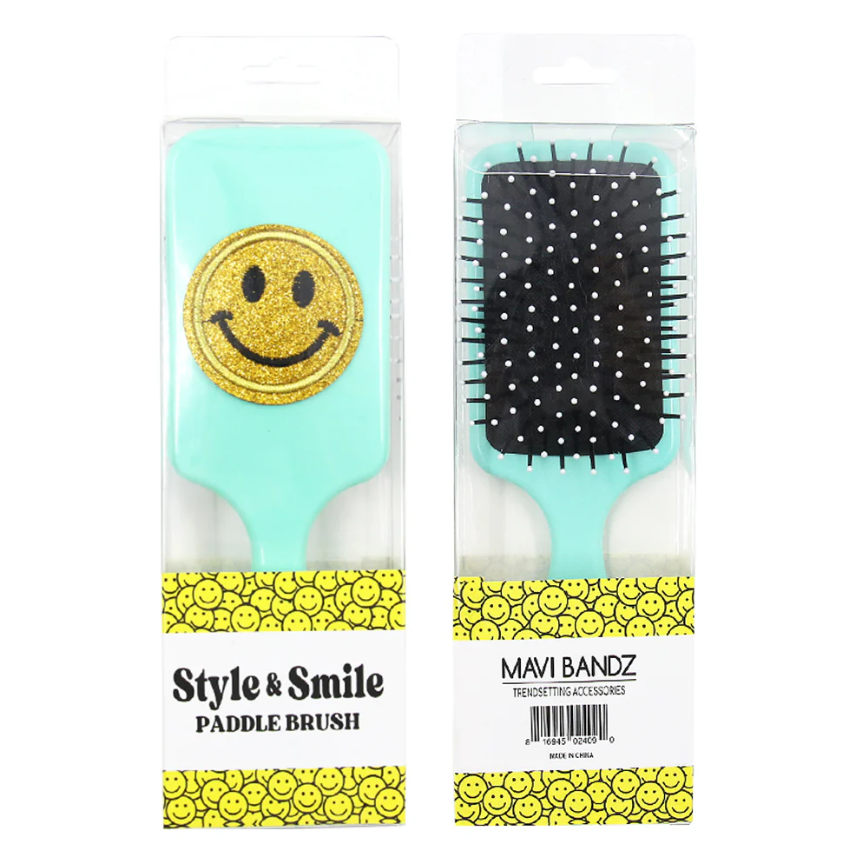 Product photography of two Mavi Bandz Style & Smile paddle brushes in clear plastic packaging, featuring light aqua handles, one with a prominent gold glitter smiley face and the other revealing a black brush pad with white polka dots, all on packaging with a vibrant yellow background patterned with small smiling faces
