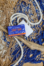 Product photograph of Gentlemen's Hardware The Fisherman's Soap Bar with its vibrant orange and blue packaging and an unwrapped white soap bar, arranged on a textured wooden surface featuring blue and tan fishing nets, thick white rope, and a piece of driftwood