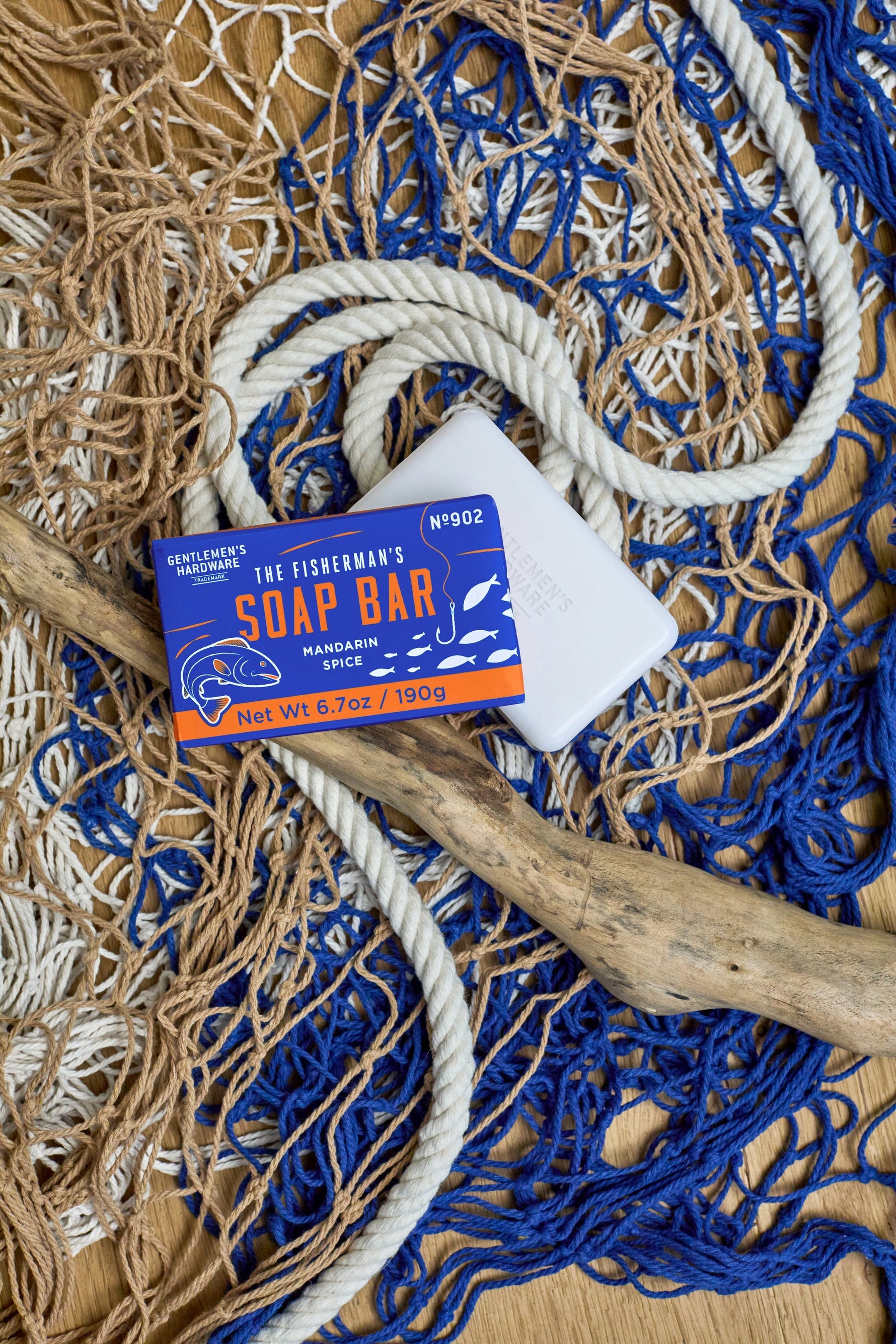 Product photograph of Gentlemen's Hardware The Fisherman's Soap Bar with its vibrant orange and blue packaging and an unwrapped white soap bar, arranged on a textured wooden surface featuring blue and tan fishing nets, thick white rope, and a piece of driftwood