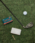 Overhead flat lay showing golf equipment including a Callaway Big Bertha iron, Nitron golf ball, and wooden tee placed next to a teal Gentlemen's Hardware Sandalwood Soap Bar box and white soap on green artificial turf