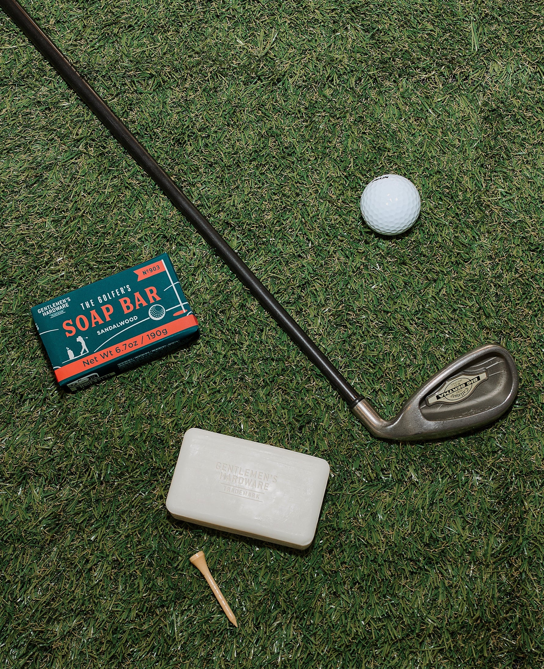 Overhead flat lay showing golf equipment including a Callaway Big Bertha iron, Nitron golf ball, and wooden tee placed next to a teal Gentlemen's Hardware Sandalwood Soap Bar box and white soap on green artificial turf