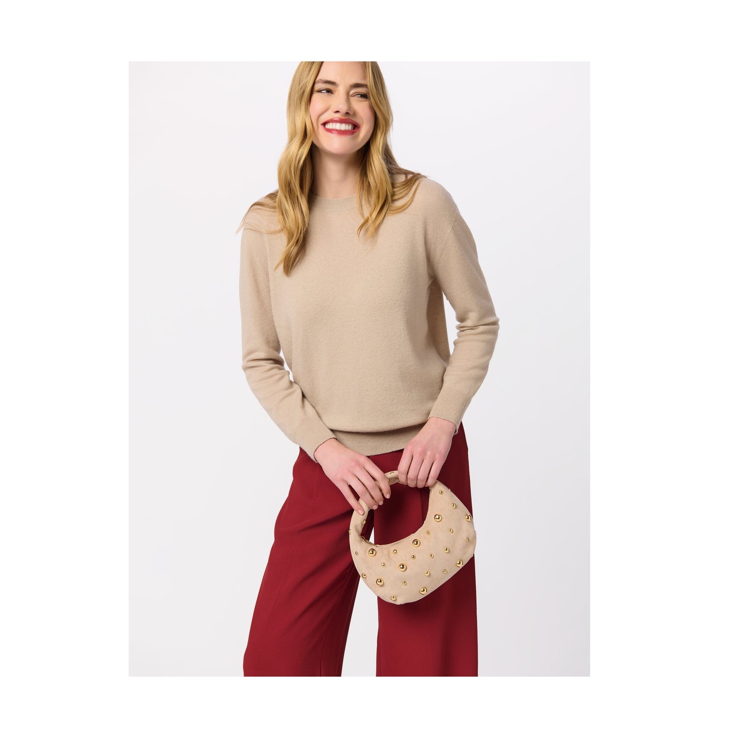 Woman wearing a beige crew neck sweater and red trousers holding a small beige studded handbag