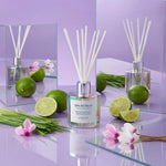 An iridescent "SPA RETREAT" diffuser bottle with white reeds is displayed on a glossy purple surface with glass reflections, surrounded by fresh green limes, bright green grass, and delicate pink cyclamen flowers