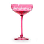 A fuchsia pink champagne coupe glass featuring ornate cut crystal circular patterns on the bowl, a slender stem, and containing a dark red liquid on a white background