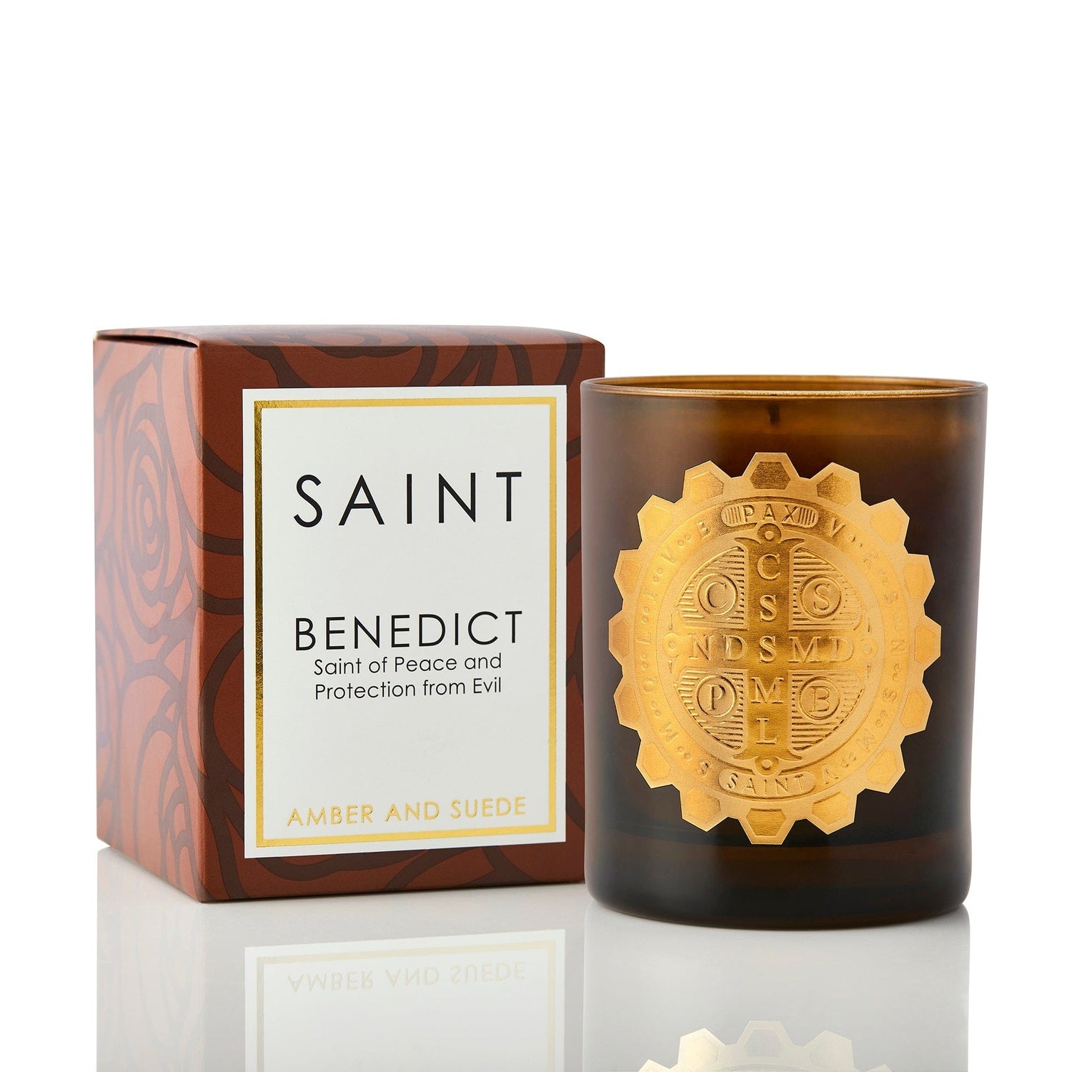 Product photograph of a Saint Benedict Amber and Suede candle in a dark amber glass jar with a prominent gold metallic Saint Benedict medal, next to its brown box displaying a subtle rose pattern and text "Saint of Peace and Protection from Evil"