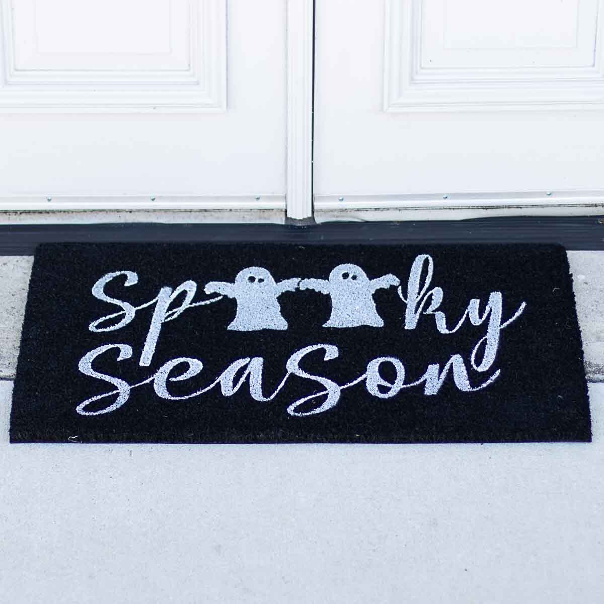 Halloween doormat at a front door featuring a black mat with white glitter Spooky Season text and two ghost figures holding hands in front of white paneled double doors