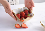 Photograph of hands holding a unique irregularly shaped gold and white bowl brimming with vibrant red strawberries, with two halved strawberries resting on a clean white marble countertop