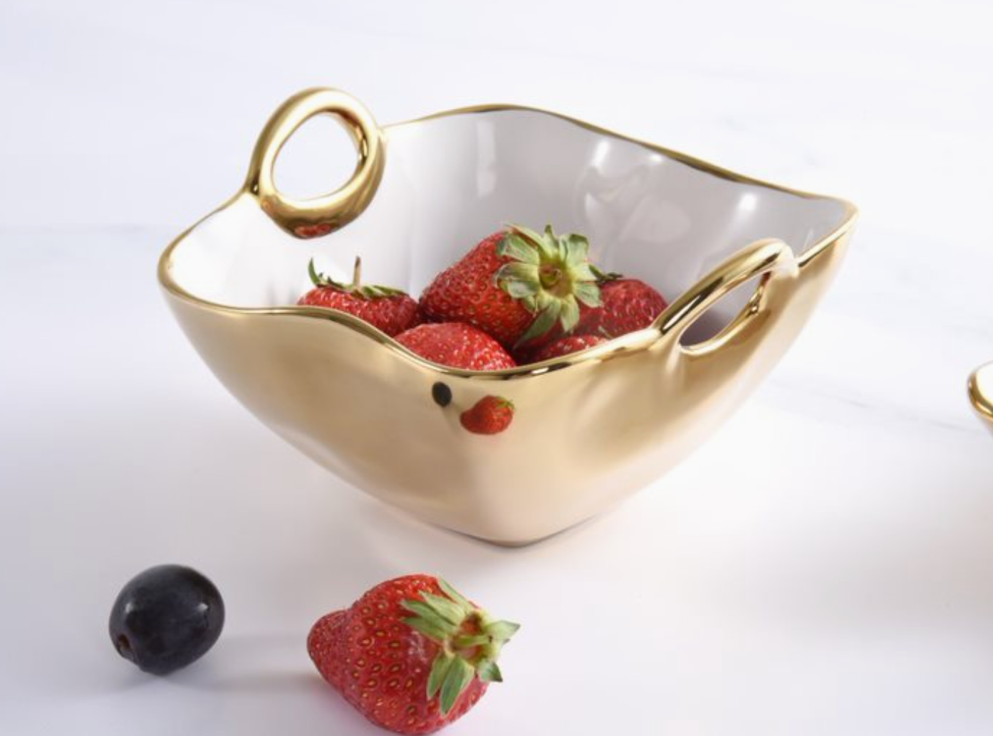 Gold and white ceramic serving bowl with two elegant handles and a wavy rim, filled with bright red strawberries, resting on a white surface with a loose strawberry and black grape
