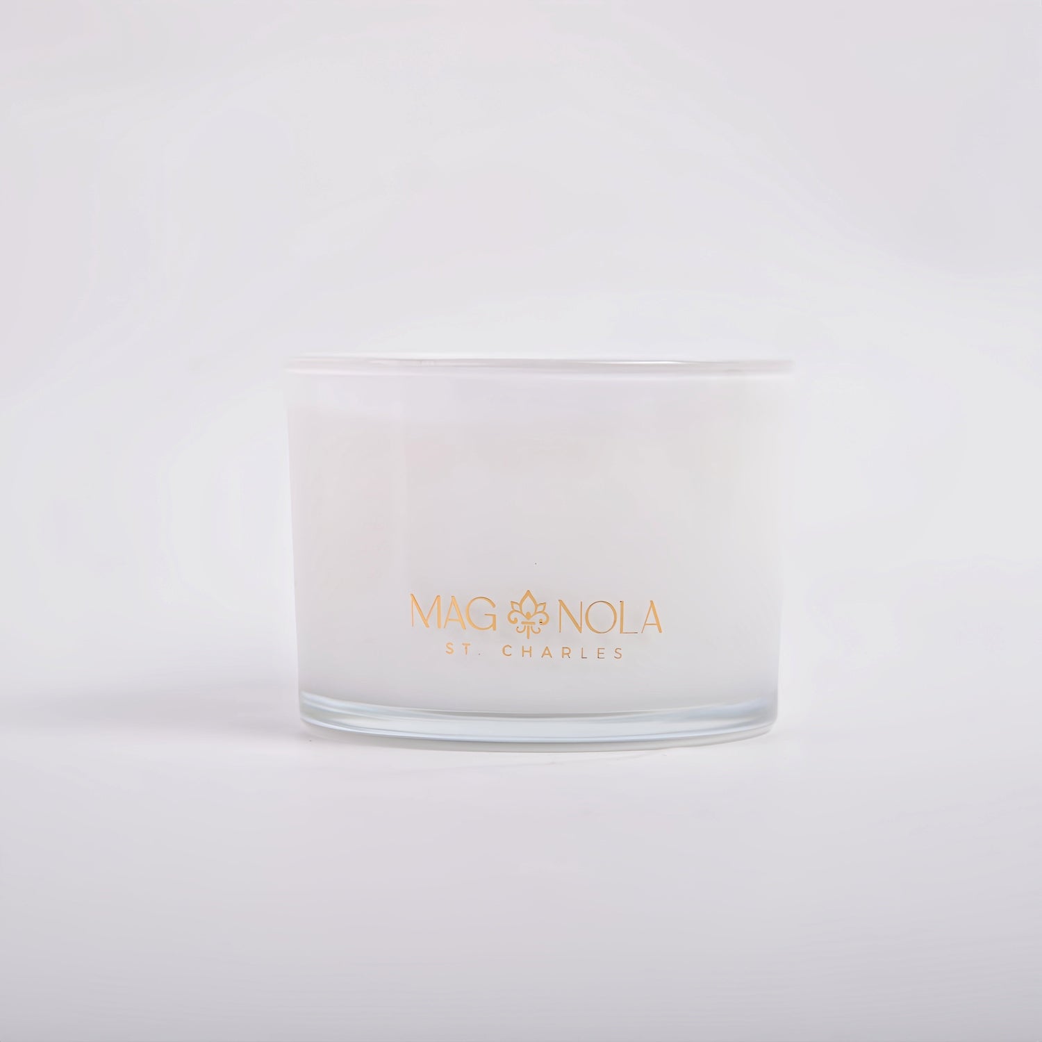 Photograph of a white candle in a frosted glass jar featuring gold "MAGNOLIA ST. CHARLES" text and a fleur-de-lis logo on a plain white background