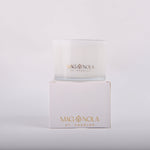 A white Magnolia St Charles candle in a glass jar sitting atop its matching white box both featuring gold foil branding with a fleur-de-lis symbol