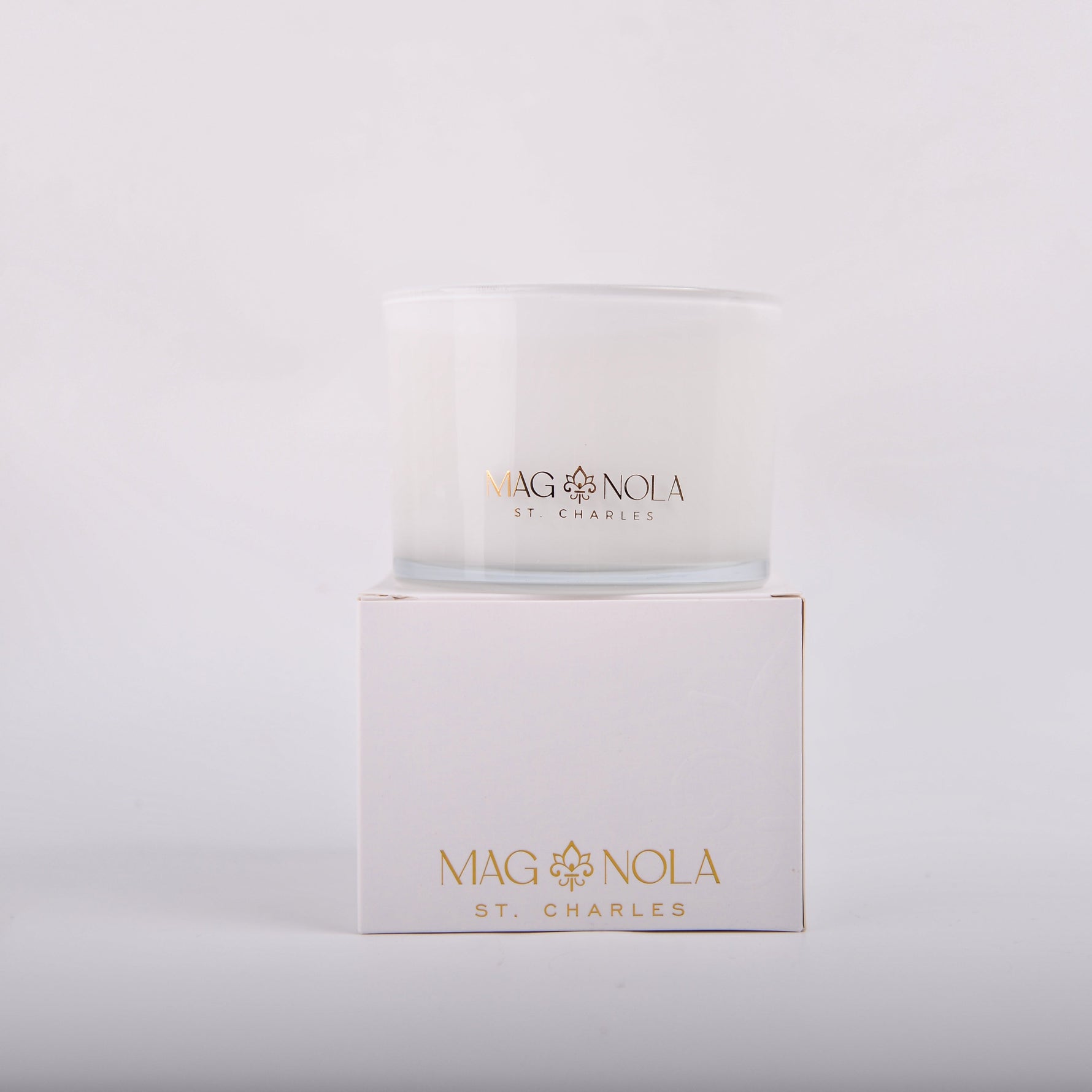 A white Magnolia St Charles candle in a glass jar sitting atop its matching white box both featuring gold foil branding with a fleur-de-lis symbol