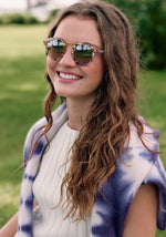 A smiling young woman outdoors wearing colorful speckled sunglasses reflecting palm trees, with a purple and white tie-dye sweater draped over her shoulders and long wavy brown hair