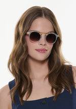 A close-up photograph of a woman with long wavy brown hair, wearing translucent light blue sunglasses with a gold bridge, and shiny pink lipstick