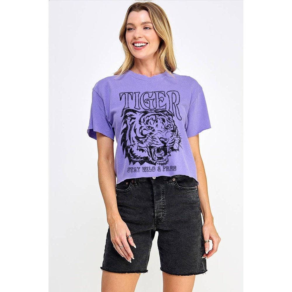 Studio fashion photograph of a smiling blonde model in a white studio wearing a purple graphic crop top with a snarling tiger print and STAY WILD & FREE text paired with black distressed denim shorts