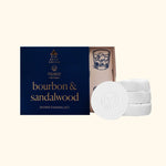 Musee -for men- bourbon & sandalwood shower steamers displayed with an open dark blue box featuring gold text and a whiskey glass illustration inside, alongside a stack of four white circular steamers each with a subtle leaf logo