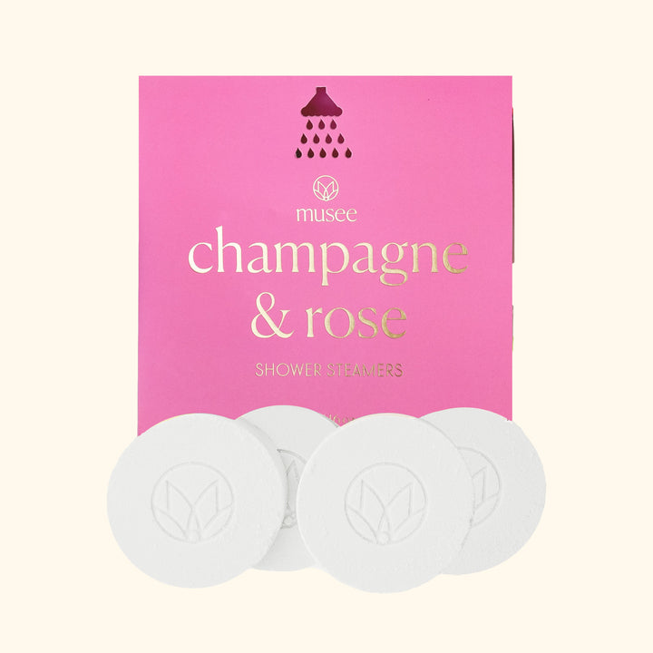 Product photography by Musee featuring Champagne & Rose Shower Steamers in vibrant pink packaging with gold foil text and a shower head icon, alongside four white round steamers embossed with a leaf logo