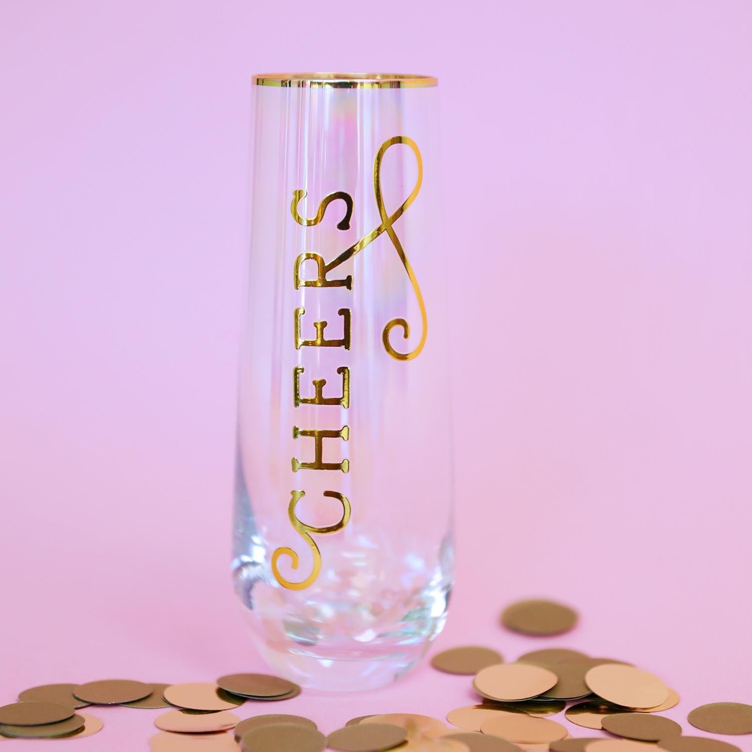 Iridescent champagne flute with gold CHEERS lettering and rim on a pink background surrounded by scattered gold confetti