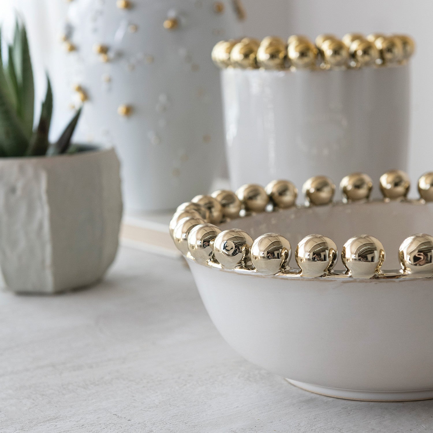 White ceramic decorative bowl with prominent shiny gold spherical rim and a taller matching vase, accompanied by a small green succulent in a geometric white pot on a light tabletop