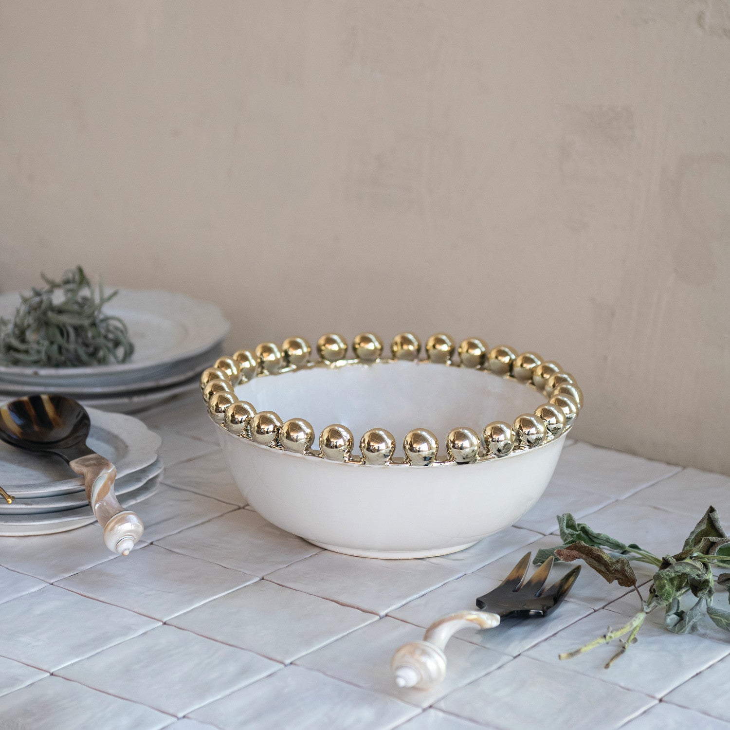 White ceramic bowl with a golden beaded rim resting on a white tiled surface beside a shell-handled serving spoon and fork, stacked white plates, and fresh green herbs