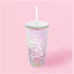 An iridescent tumbler cup featuring white script spelling "bride" with floating pink and purple glitter inside, topped with a white lid and straw against a plain pink background