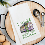 White kitchen towel featuring a purple and green New Orleans streetcar decorated with Mardi Gras beads and a fleur-de-lis, with the green text LAISSEZ LES BON TEMPS ROULER, resting on a wooden tray alongside measuring spoons
