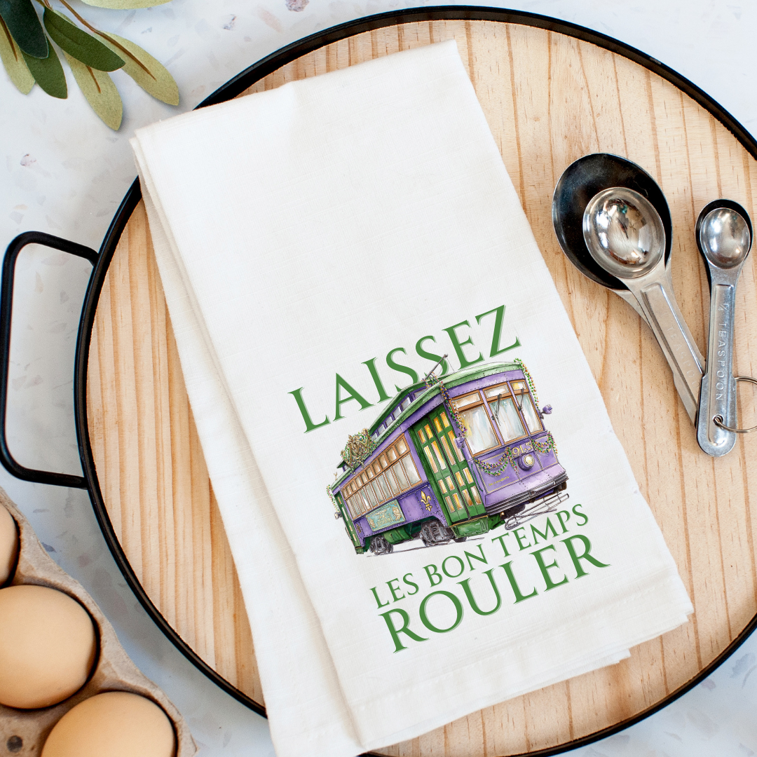 White kitchen towel featuring a purple and green New Orleans streetcar decorated with Mardi Gras beads and a fleur-de-lis, with the green text LAISSEZ LES BON TEMPS ROULER, resting on a wooden tray alongside measuring spoons