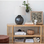 A modern interior features a light wood console table with a slatted design, displaying a black and silver mother-of-pearl mosaic lamp, a framed abstract artwork with swirling beige lines, and a hanging green plant in a textured grey pot