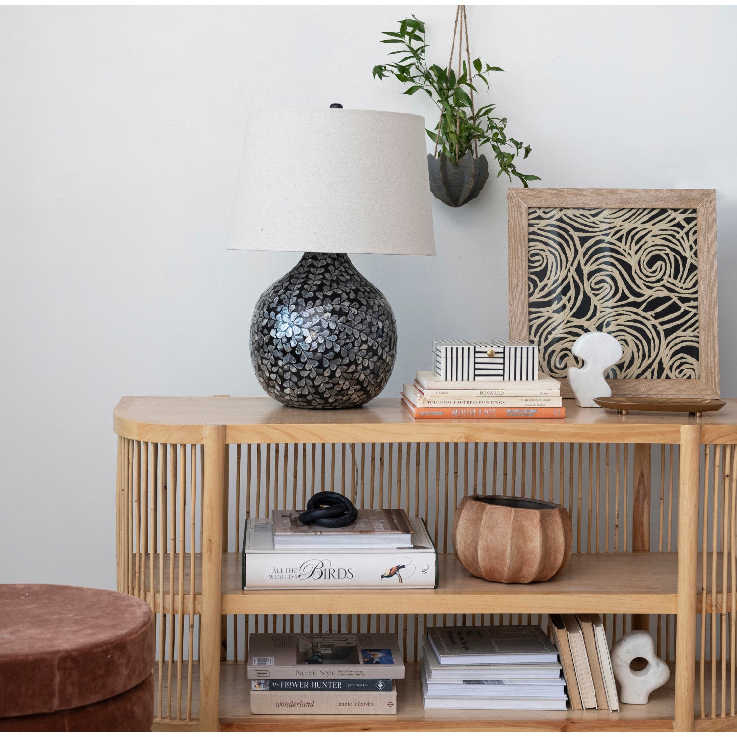 A modern interior features a light wood console table with a slatted design, displaying a black and silver mother-of-pearl mosaic lamp, a framed abstract artwork with swirling beige lines, and a hanging green plant in a textured grey pot