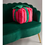 A plush gift-shaped pillow in vibrant pink and red stripes with a green velvet bow rests on a luxurious emerald green velvet sofa featuring a distinctive scalloped back and golden metal leg