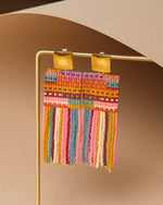 Handcrafted multi-colored seed bead fringe earrings featuring square gold studs and a vibrant geometric woven top with dangling strands in pink, blue, yellow, and orange, displayed on a gold stand against a neutral background