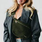 A smiling blonde woman wearing a grey pinstripe blazer and an olive green woven crossbody bag with gold hardware and three small studs
