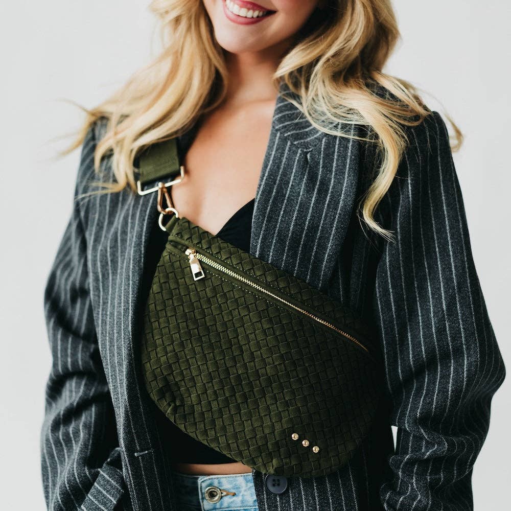 A smiling blonde woman wearing a grey pinstripe blazer and an olive green woven crossbody bag with gold hardware and three small studs