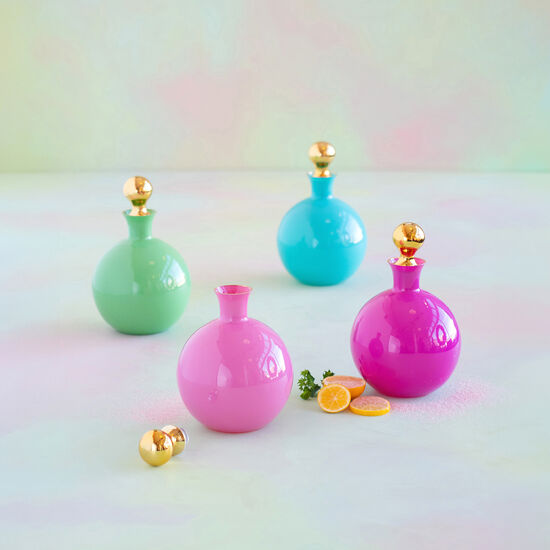 Photograph of four glossy spherical bottles in bright green, turquoise, and two pink shades, featuring gold stoppers, on a soft pastel background with scattered lemon slices and loose gold spheres