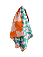 Textile by crae a vibrant folded towel showcasing an orange and light pink organic pattern on one side and an emerald green and light pink curved checkerboard design on the reverse with a textured waffle weave