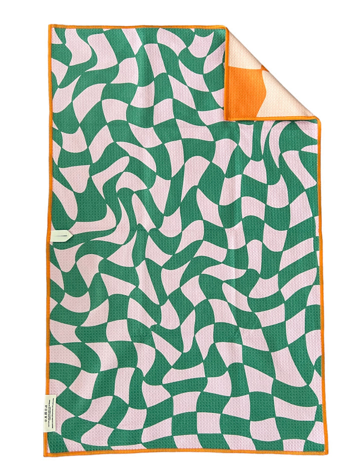 Towel with a playful warped emerald green and light pink checkerboard pattern featuring an orange trim and a corner folded to reveal its solid orange reverse