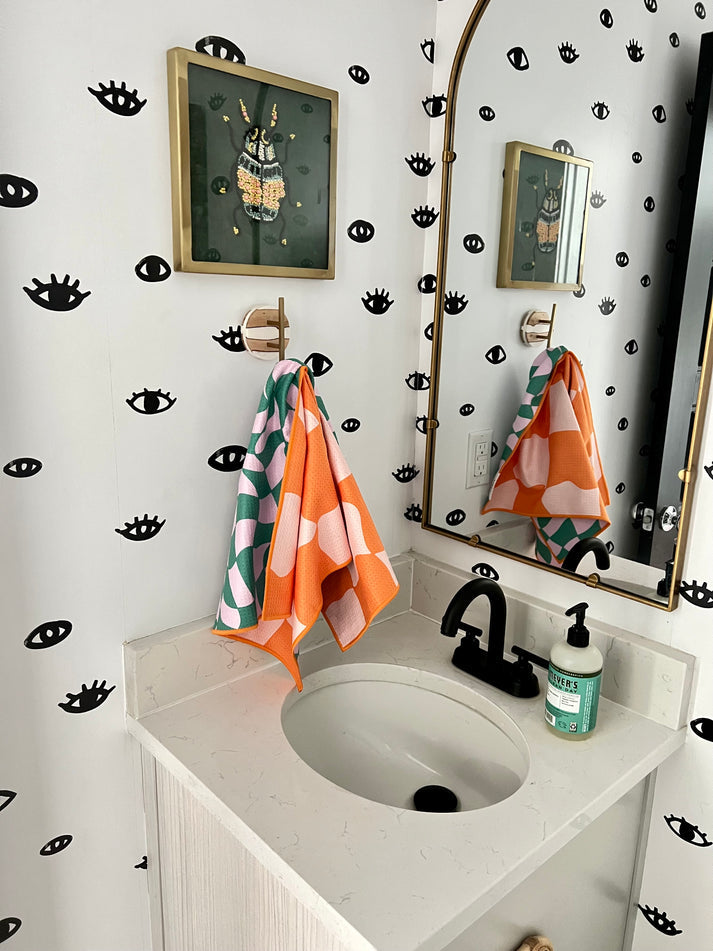 A modern bathroom vanity featuring white walls with black eye decals, a gold-framed beaded beetle artwork above two colorful green and orange patterned hand towels, reflected in a brass arched mirror
