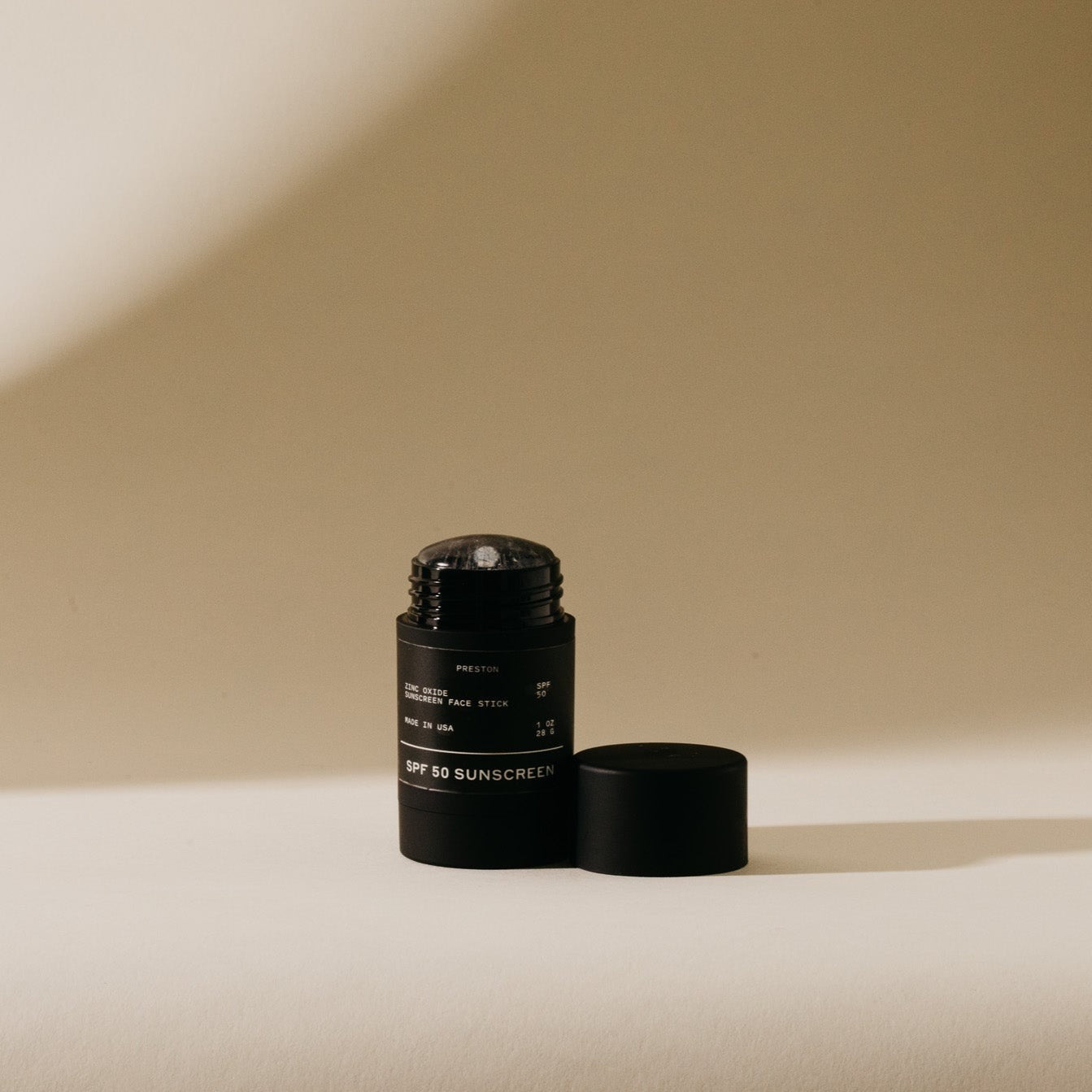 Photograph of an uncapped black Preston Zinc Oxide SPF 50 Sunscreen Face Stick with white lettering and a matching cap beside it on a bright surface against a warm beige background with a diagonal shadow