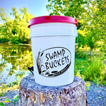White "Swamp Buckets" bucket with a bright red lid, a black logo featuring cattails and a water lily, and a small blue spigot, resting on a textured tree stump by a sunlit pond in Brusly, Louisiana
