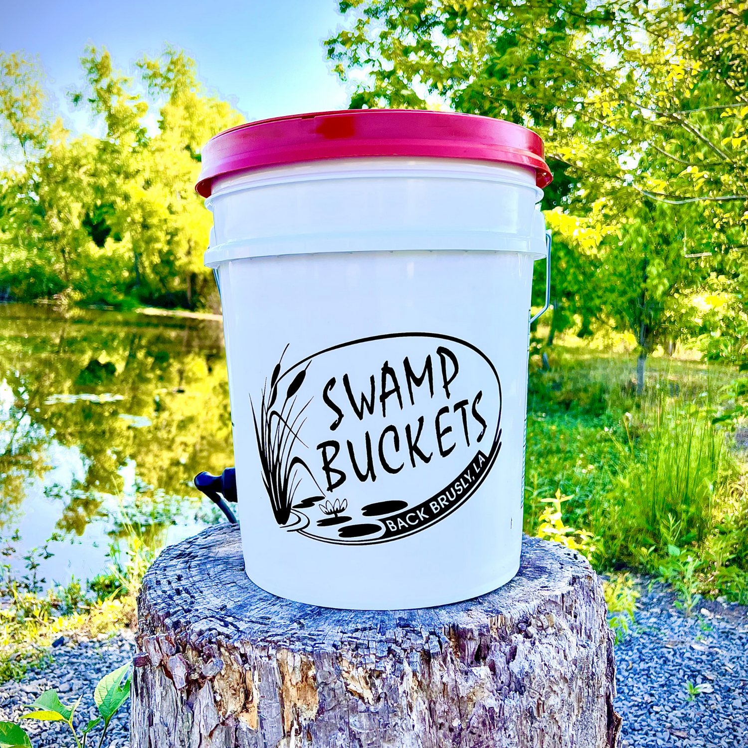 White "Swamp Buckets" bucket with a bright red lid, a black logo featuring cattails and a water lily, and a small blue spigot, resting on a textured tree stump by a sunlit pond in Brusly, Louisiana