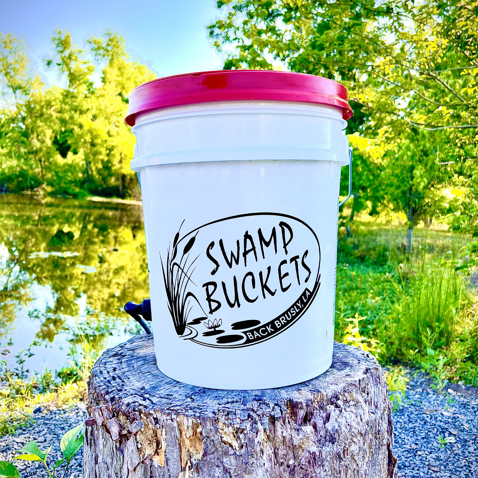 White "Swamp Buckets" bucket with a bright red lid, a black logo featuring cattails and a water lily, and a small blue spigot, resting on a textured tree stump by a sunlit pond in Brusly, Louisiana
