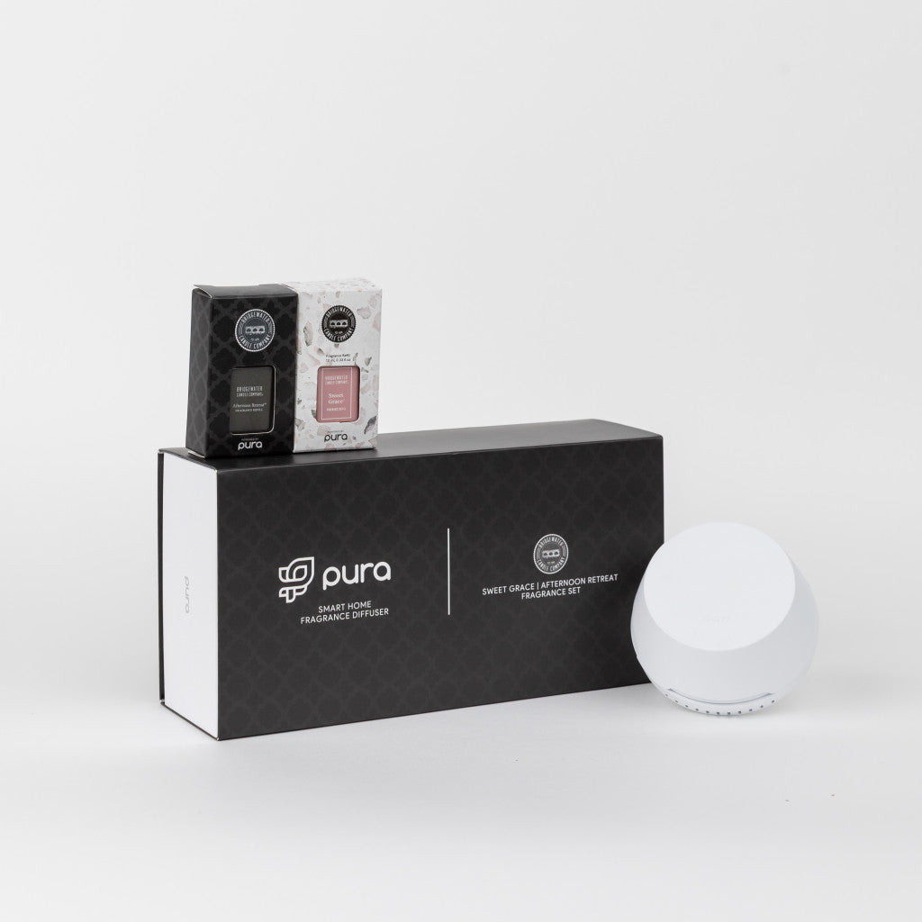 Photograph of a Pura smart home fragrance diffuser set on a white studio background, showcasing a black Pura gift box prominently labeled 'Smart Home Fragrance Diffuser', a white Pura smart diffuser device, and two smaller fragrance refill boxes, one black Brightwater and one pink Sweet Grace