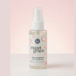 Photograph of a clear Sweet Grace Stain Remover spray bottle with a white pump top, featuring a white label with 'sweet grace' text and light pink and orange concentric circle patterns, set against a soft pink gradient background