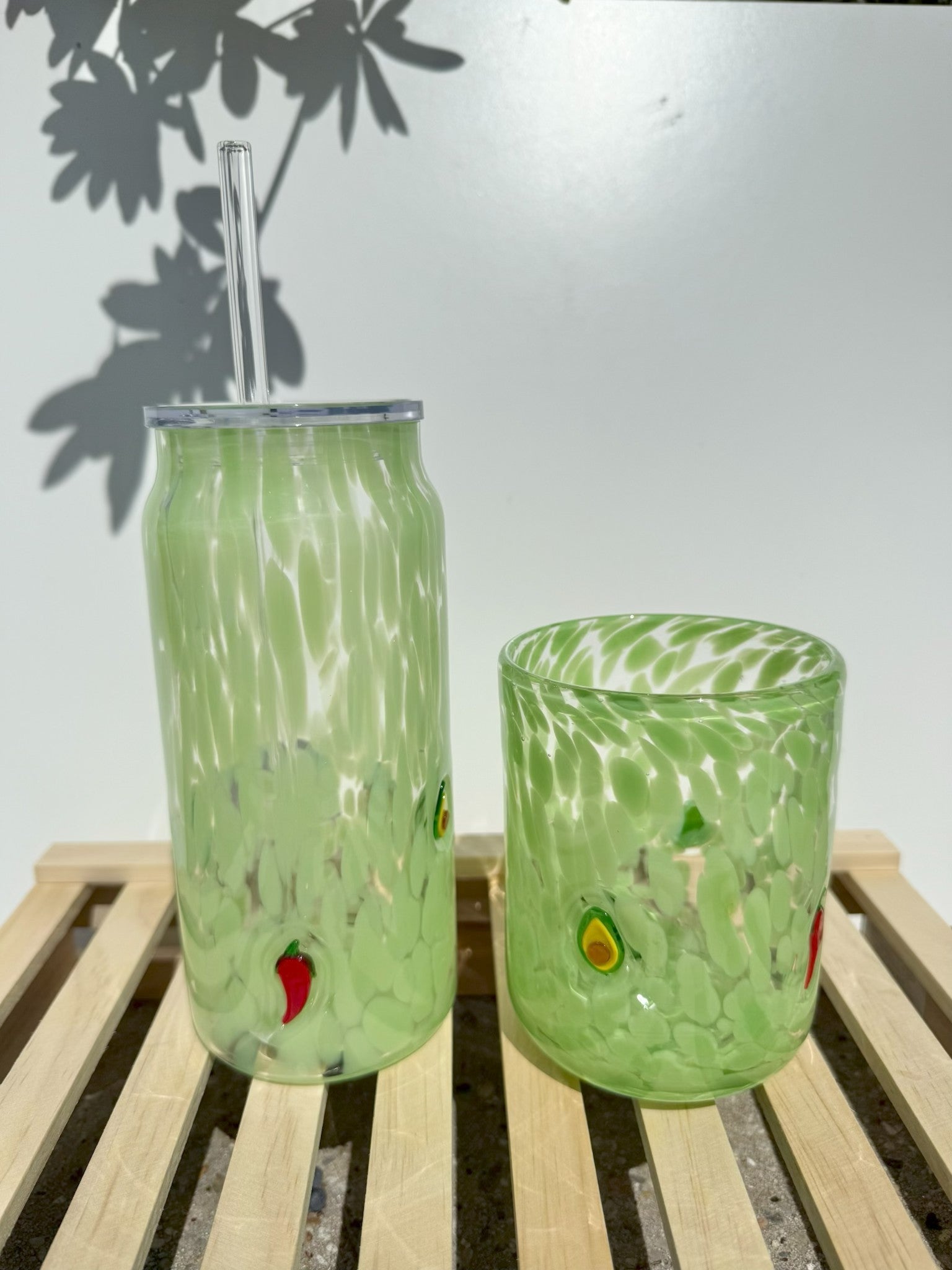 Handblown glass avocado and chili pepper themed drinking glasses on a wooden slat surface against a white wall with dappled plant shadows, featuring a tall tumbler with a clear straw and a shorter wide glass, both speckled light green with embedded red chili peppers and green and yellow avocado slices